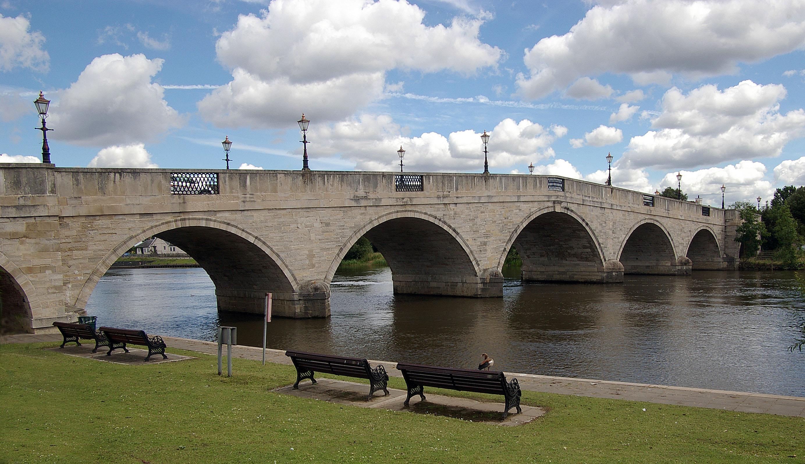 Chertsey Bridge, the elegant 18th-century stone arch bridge spanning the River Thames at Chertsey, Surrey, with riverside benches and a bright blue sky