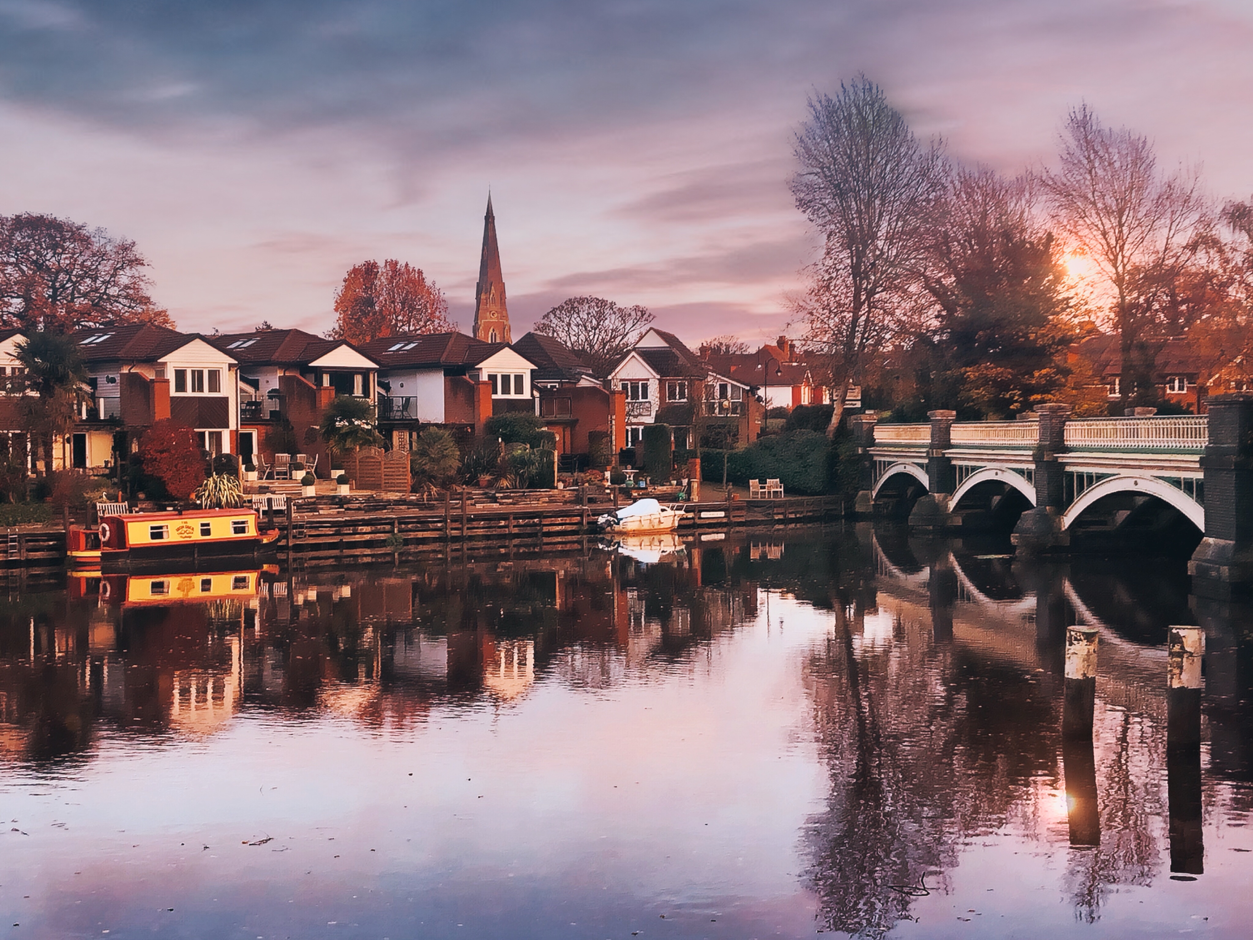 Golden-hour view of the River Wey at Weybridge, Surrey, with a narrowboat moored along the bank, St James's church spire rising above the riverside houses, and the old bridge reflected in the still water at sunset